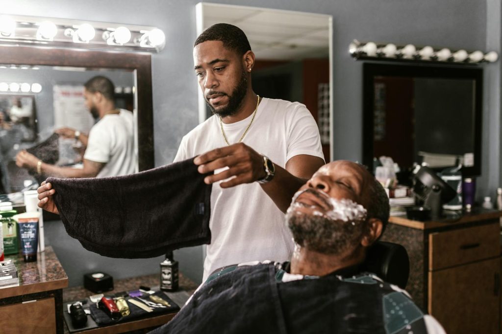 Barber prepares to shave a man with a towel in a modern barbershop.